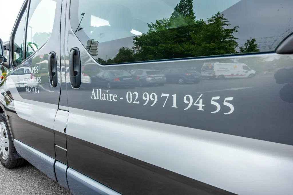 Flanc de véhicule corbillard gris de pompes funèbres avec numéro de téléphone.